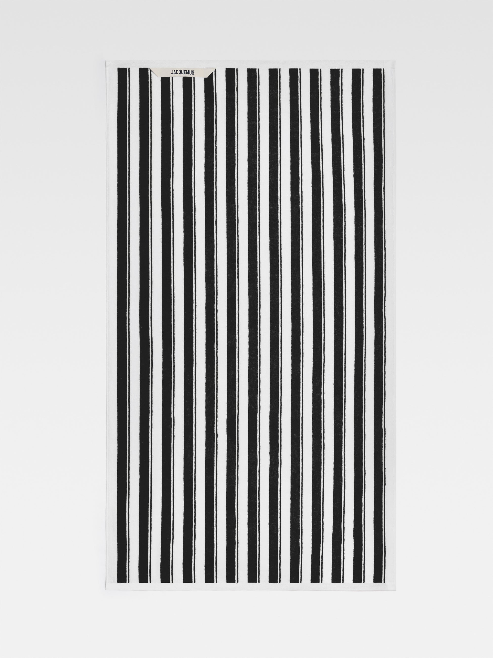 A close-up view of a large beach towel with bold black and white stripes. The Jacquemus logo is visible in the top left corner.