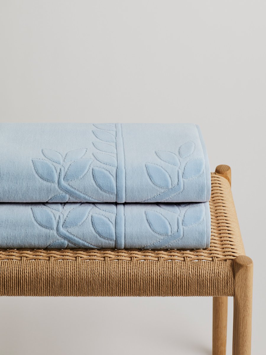 Two 'Maldivian Atoll' beach towels folded on a wooden bench. The towels feature a subtle leaf pattern.