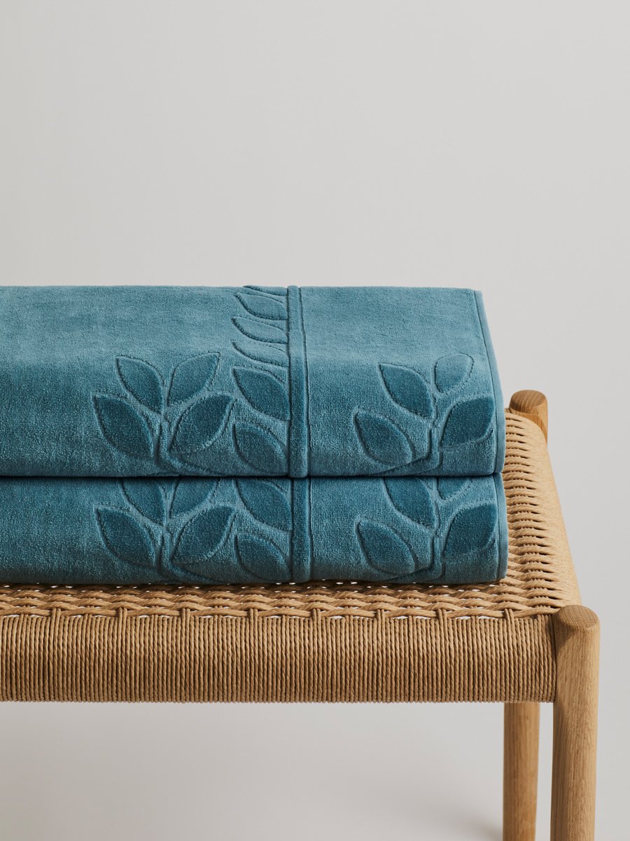 Two 'Polynesian Lagoon' beach towels folded on a wooden bench. The towels feature a subtle leaf pattern.
