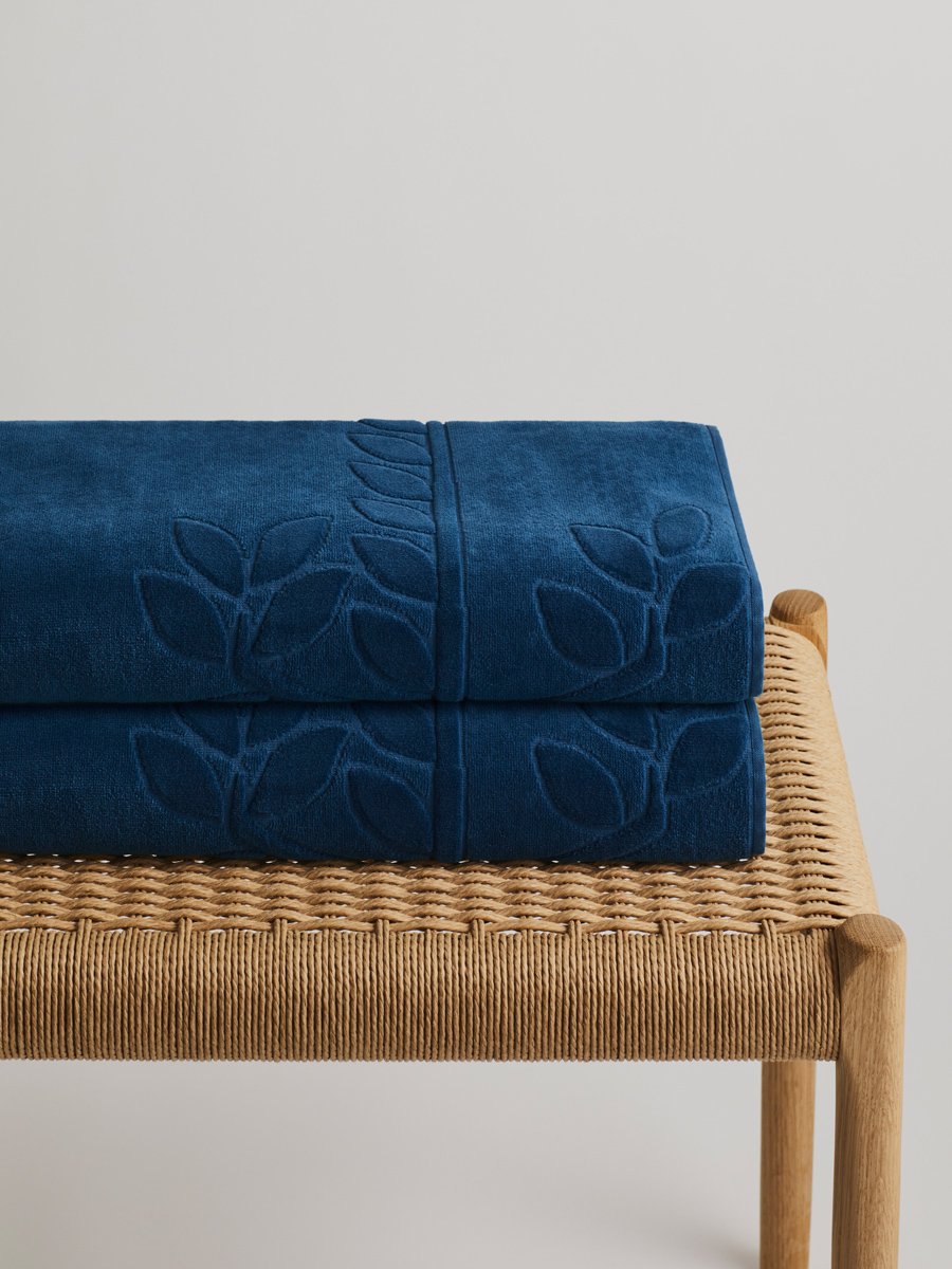 Two 'Riviera Navy' beach towels folded on a wooden bench. The towels feature a subtle leaf pattern.