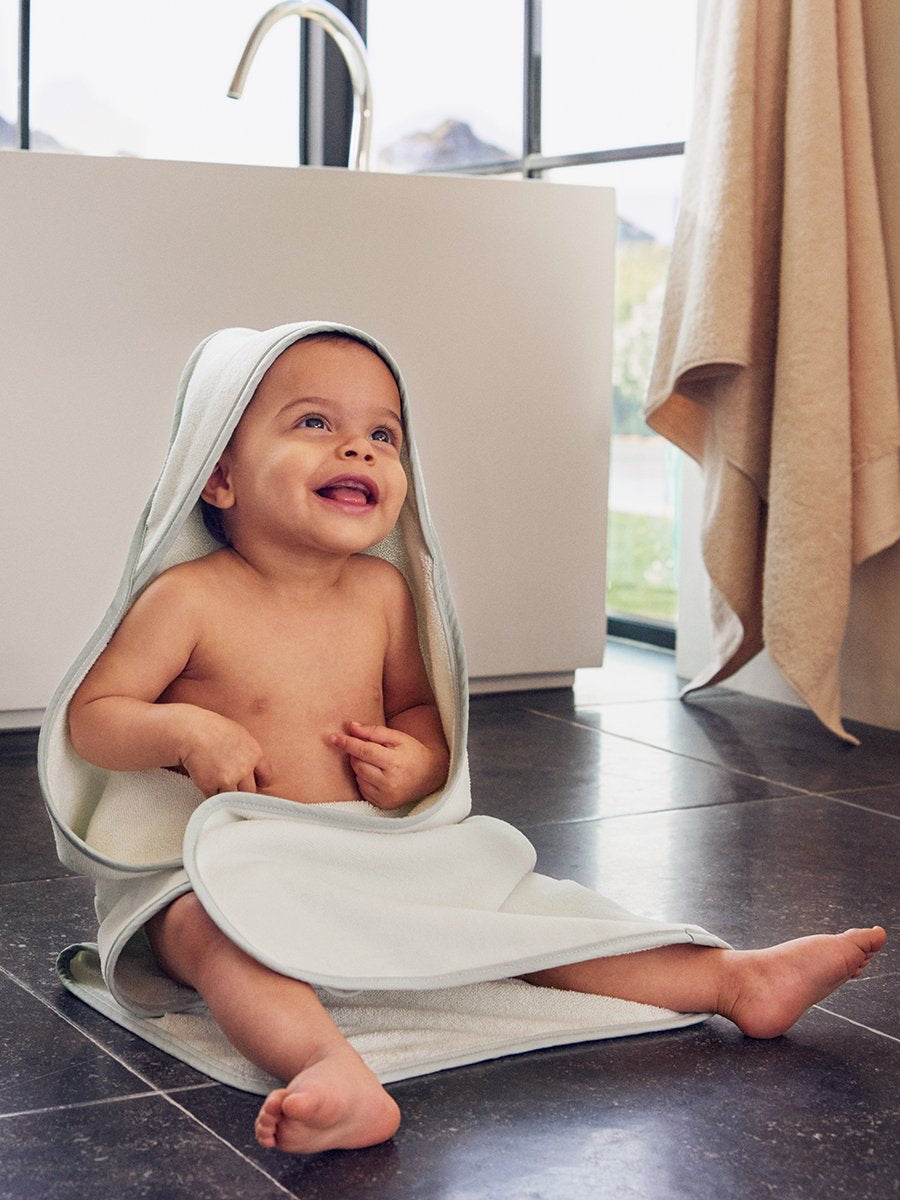 A baby wearing a hooded towel, smiling while sitting near a bathtub.