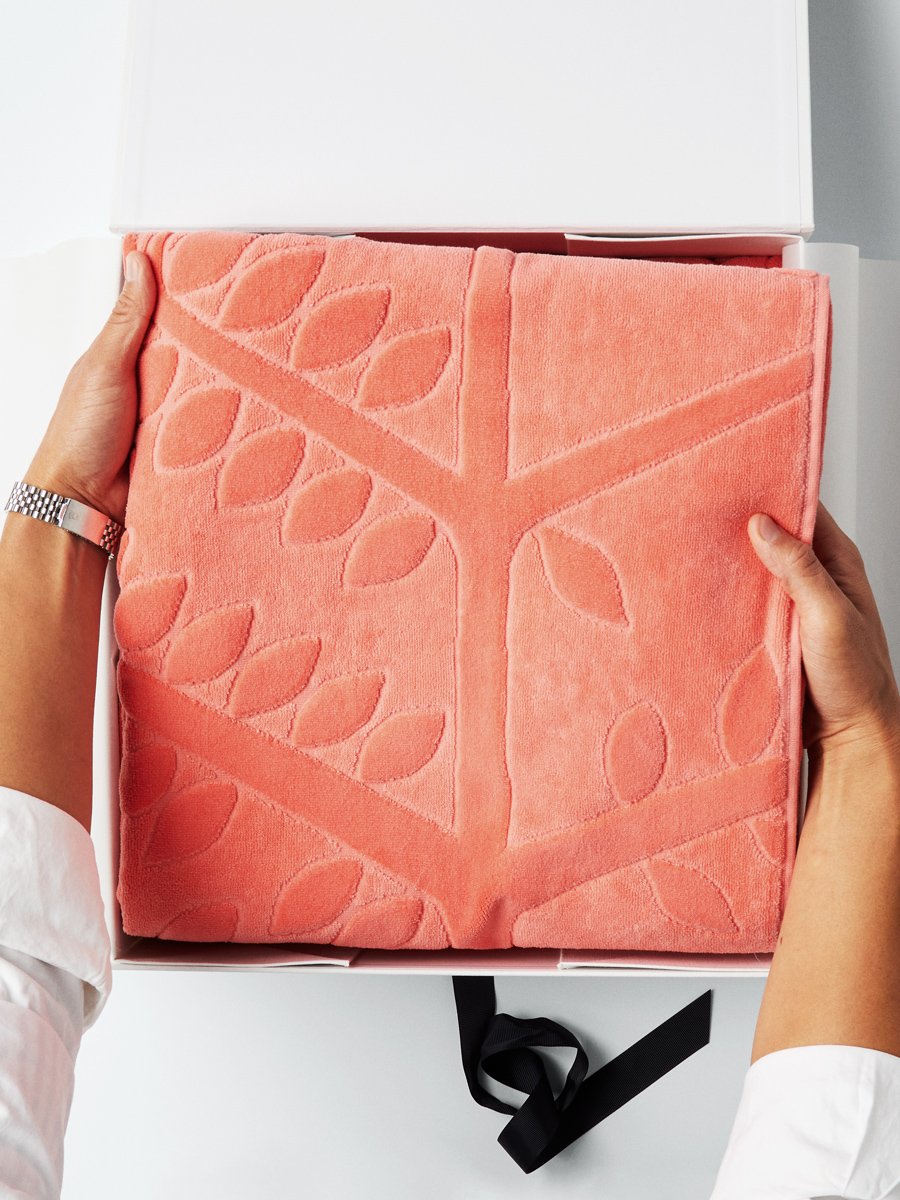A pair of hands holding a folded beach towel with a FourSeasons logo design. The towel rests in a white box with a black ribbon.