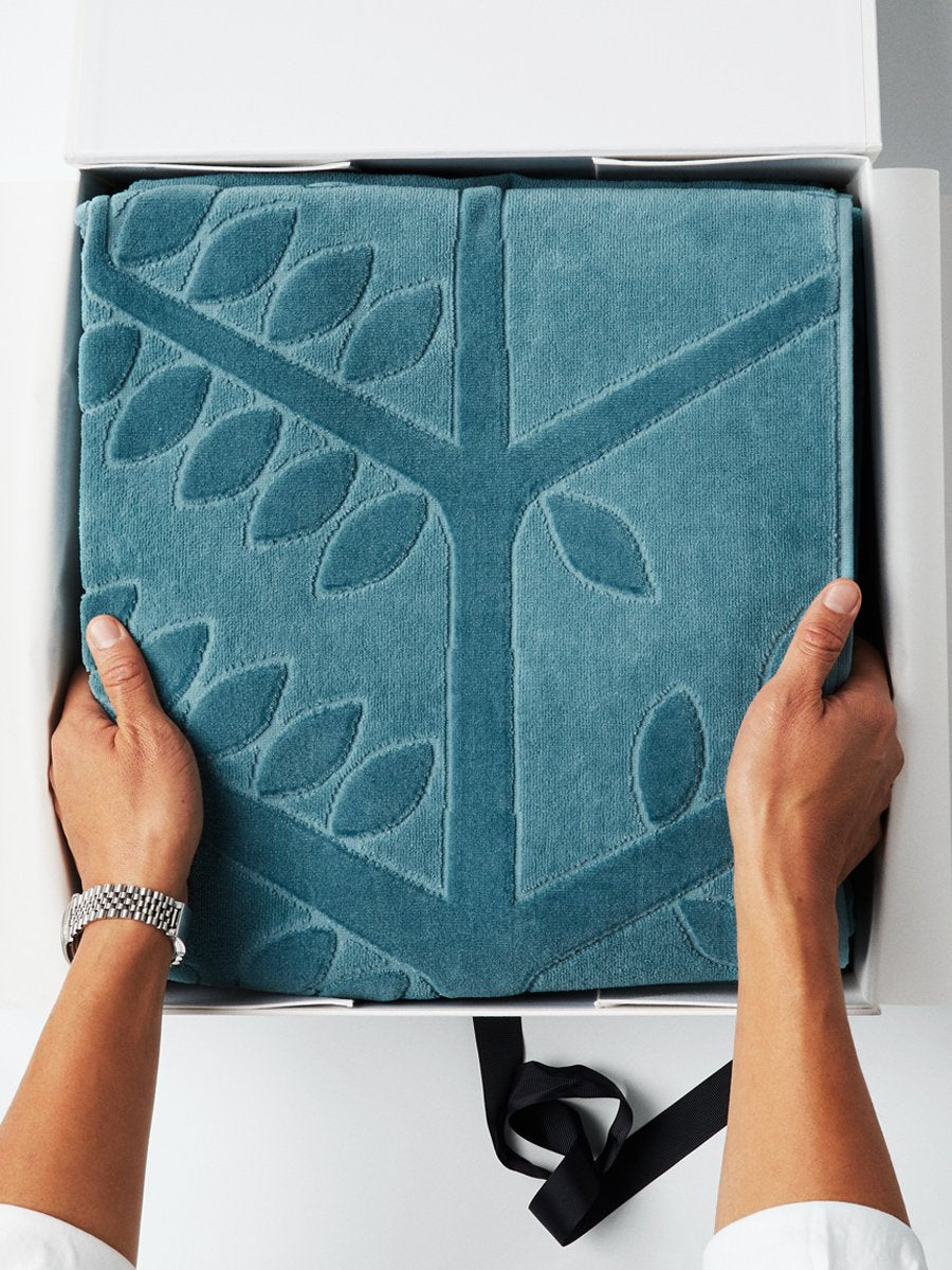 A pair of hands holding a folded beach towel with a FourSeasons logo design. The towel rests in a white box with a black ribbon.