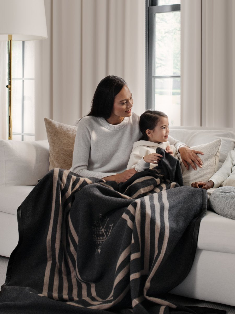 A heartwarming scene of a mother and daughter sharing a cozy moment, both wrapped in a luxurious black throw with a leather trim.