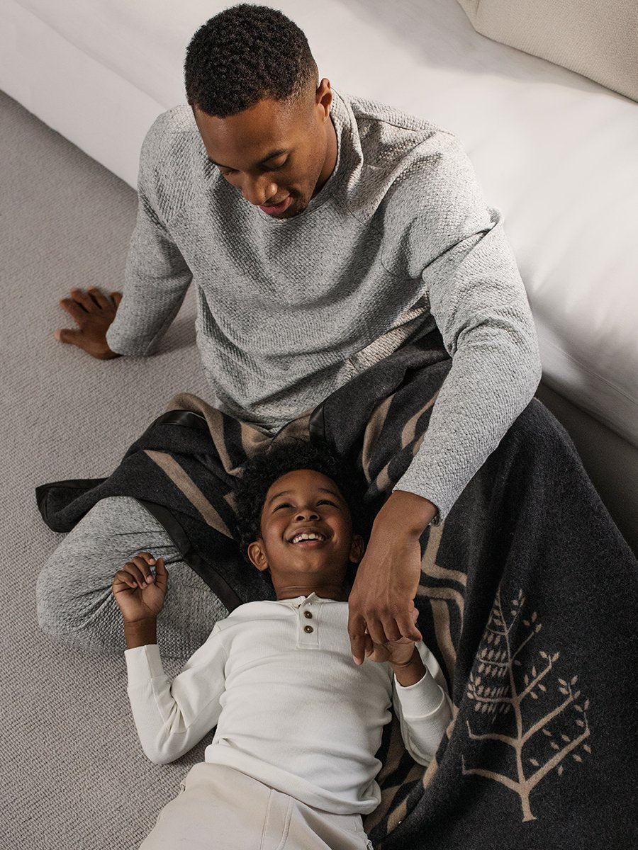 A father and son cuddling together, with the child resting on the father's lap, both covered by a black throw with a leather trim.