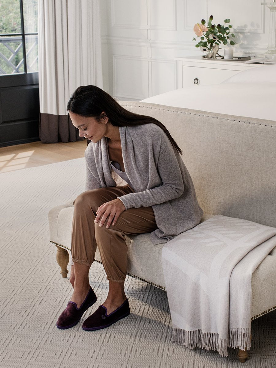 A woman sits on a chair putting on a pair of Four Seasons Velvet Slippers