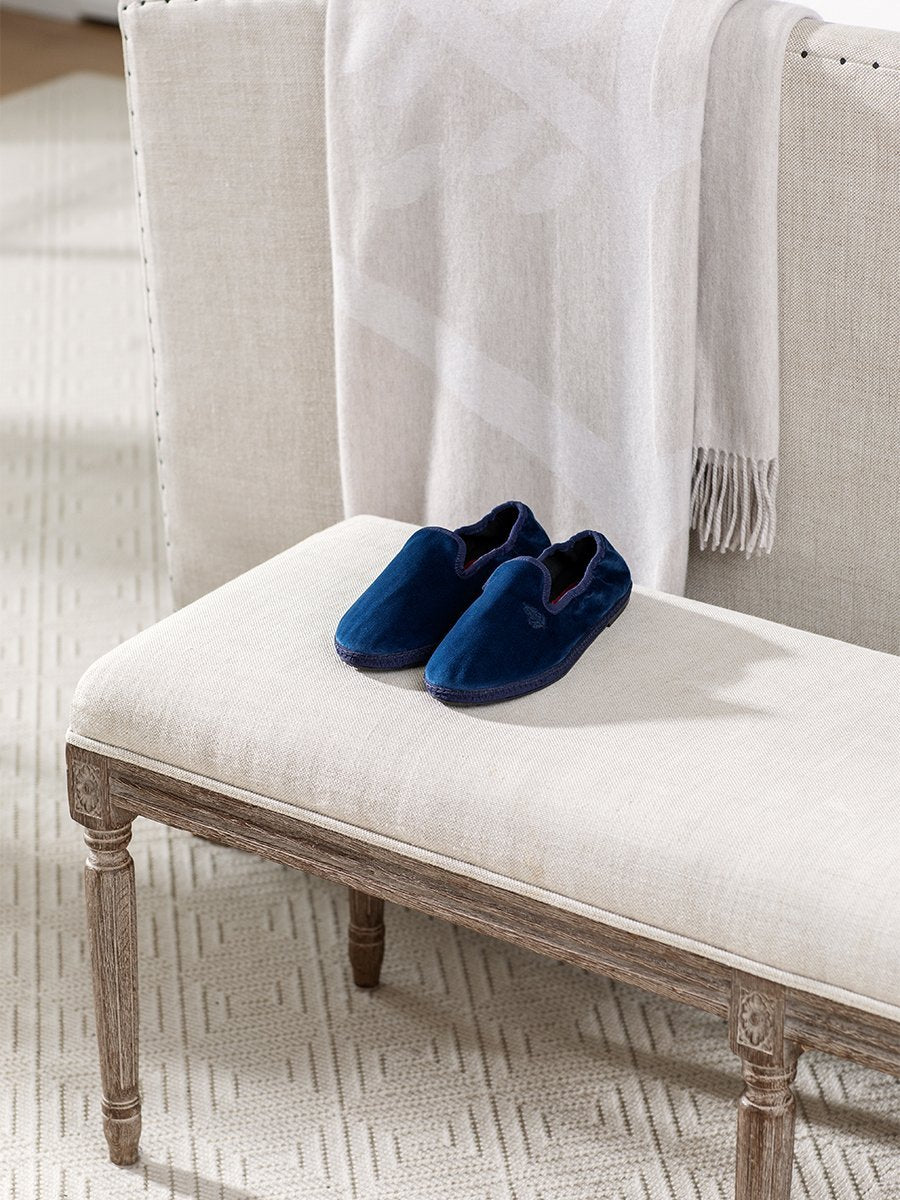 Four Seasons Velvet Slippers in Riviera Navy placed on a bench in a cozy living room setting