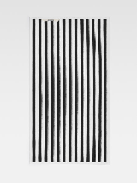 A close-up view of a large beach towel with bold black and white stripes. The Jacquemus logo is visible in the top left corner.