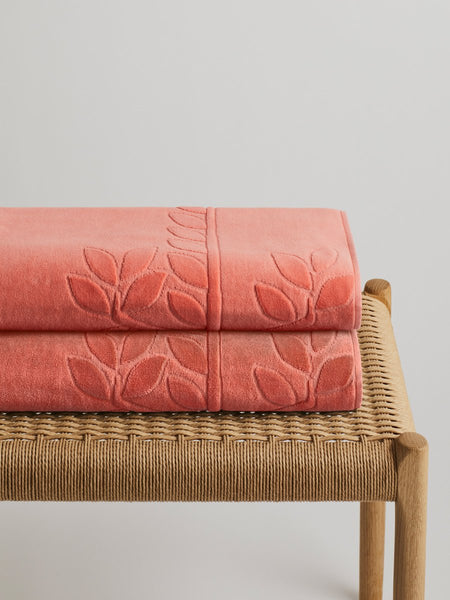 Two 'Red Sea Coral' beach towels folded on a wooden bench. The towels feature a subtle leaf pattern.