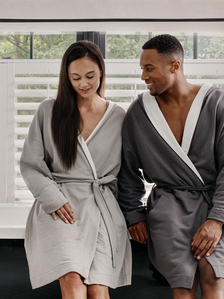 Two figures, a man and a woman, relaxing near a bathtub, dressed in white hooded robes.